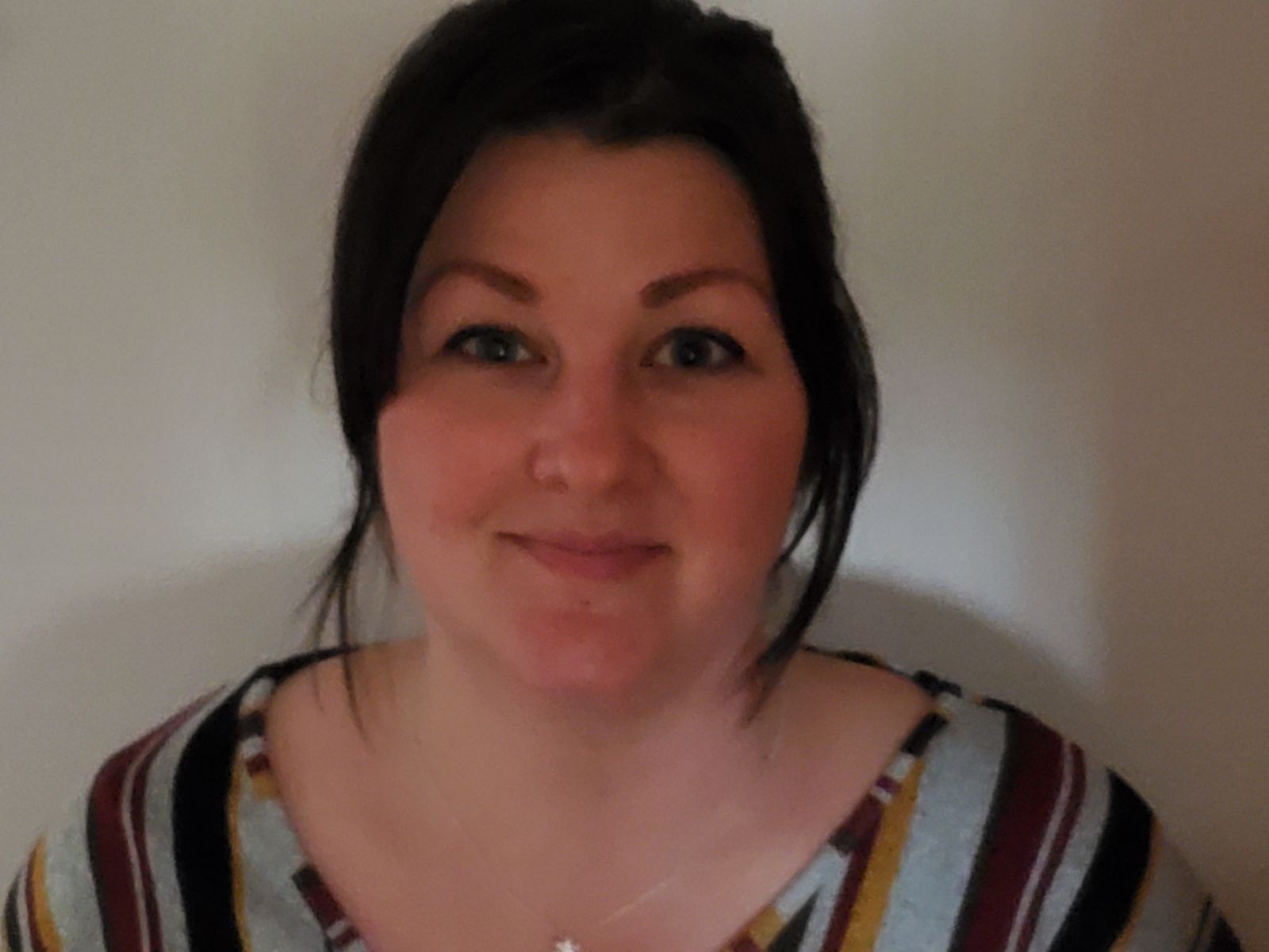 Person wearing a striped shirt with their hair tied back, standing against a plain wall.